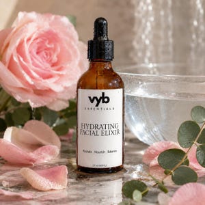 Puede incluir: Una botella de vidrio marrón de elixir facial hidratante con un cuentagotas negro, etiquetada como "vyb essentials". La etiqueta dice "Hydrating Facial Elixir" y "Hydrate. Nourish. Balance." La botella es de 60 ml. Pétalos de rosa rosas y una rosa en el fondo.