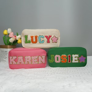 Könnte beinhalten: Drei Reißverschlusstaschen mit verschiedenen Namen. Die Taschen sind aus weichem Stoff und haben einen flauschigen, bunten Schriftzug. Die Namen sind "LUCY", "KAREN" und "JOSIE".
