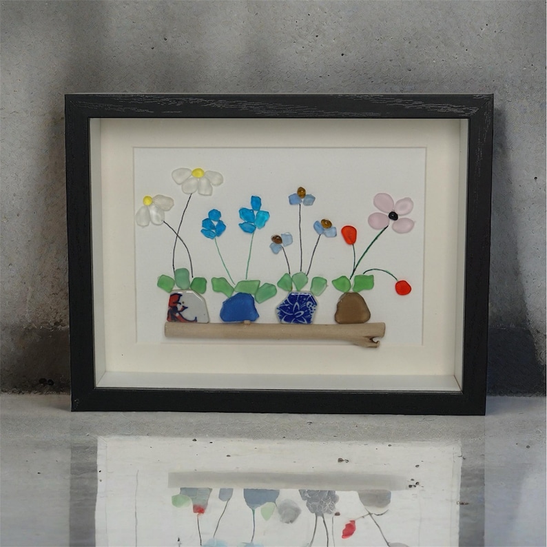 May include: A framed artwork featuring a colorful flower arrangement made of sea glass. The flowers are in various shades of blue, pink, red, and white, and are arranged in a row on a piece of driftwood. The artwork is set against a white background and is framed in a black frame.