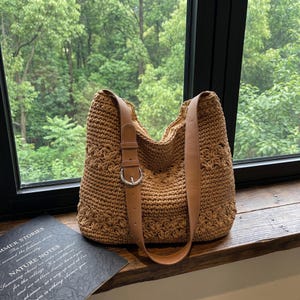 Könnte beinhalten: Eine braune, gewebte Tragetasche mit Lederriemen und Schnalle. Die Tasche hat ein Blumenmuster und einen langen Schulterriemen. Die Tasche steht auf einer Holzoberfläche in der Nähe eines Fensters.