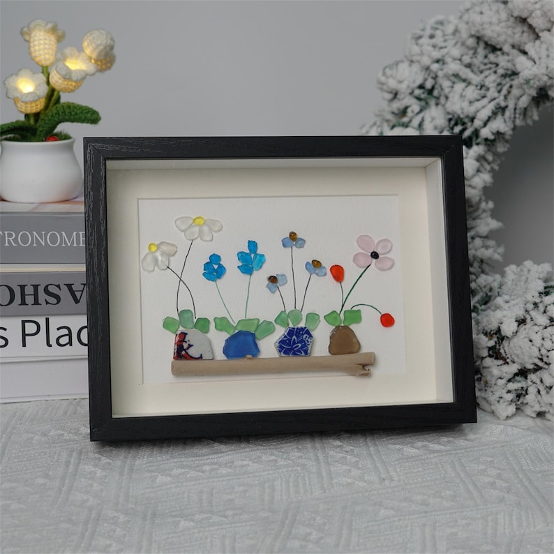 May include: Framed artwork featuring a flower arrangement made of colorful glass pieces. The flowers are arranged in a row on a wooden branch, with each flower having a different color and shape. The artwork is set against a white background and framed in black.