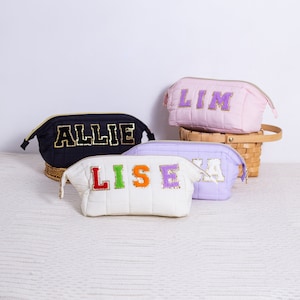 May include: Four quilted makeup bags with personalised names in colourful letters. The bags are in black, white, pink and purple.