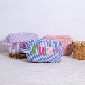 May include: Three striped fabric zippered pouches with different names embroidered on them. The names are "FUG", "JUAN", and "N".