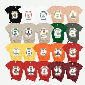 May include: A collection of t-shirts in various colors, each featuring a graphic resembling a sauce bottle label. The labels display different sauce names like "Queso Dip," "Hot Sauce," and "Chipotle Sauce." The shirts are arranged on a white background.