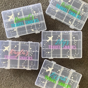 May include: Four clear plastic containers with lids, each labelled with a name and "Travel Snacks". The names are Daniel's, Kayla's, Lily's, and Ezah's. Each container has a small airplane graphic and a dashed line design.