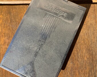 May include: Silver-toned rectangular case with the word "Bridge" in cursive. The case has a textured, cross-hatched pattern and a geometric design. The case is closed and appears to be made of metal. The case is resting on a wooden surface.