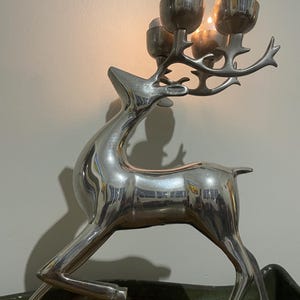 May include: A silver-toned metal reindeer candle holder featuring a polished finish. The deer is depicted in a leaping pose with its antlers extending upward to support three circular candle cups, one of which holds a lit tea light candle.