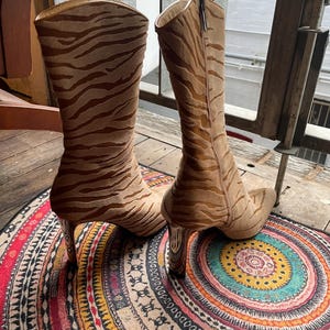 May include: A pair of tall, tan boots with a zebra stripe pattern. The boots have a pointed toe and a high, clear heel. A zipper runs up the side of each boot. The boots are displayed on a colorful, patterned rug.