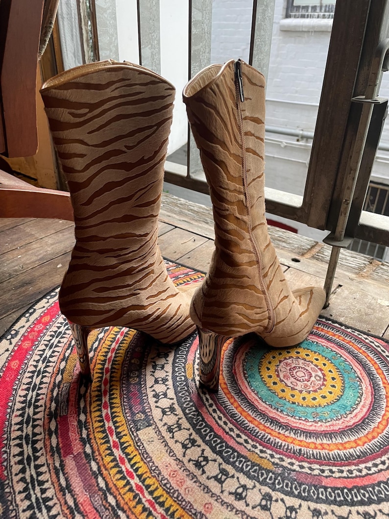 May include: A pair of tall, tan boots with a zebra stripe pattern. The boots have a pointed toe and a high, clear heel. A zipper runs up the side of each boot. The boots are displayed on a colorful, patterned rug.
