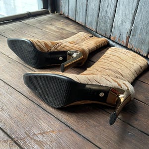 May include: A pair of tan and brown patterned high-heeled boots with gold-colored heels. The boots have a pointed toe and a zipper closure. The soles are black. The boots are made of a textured material.