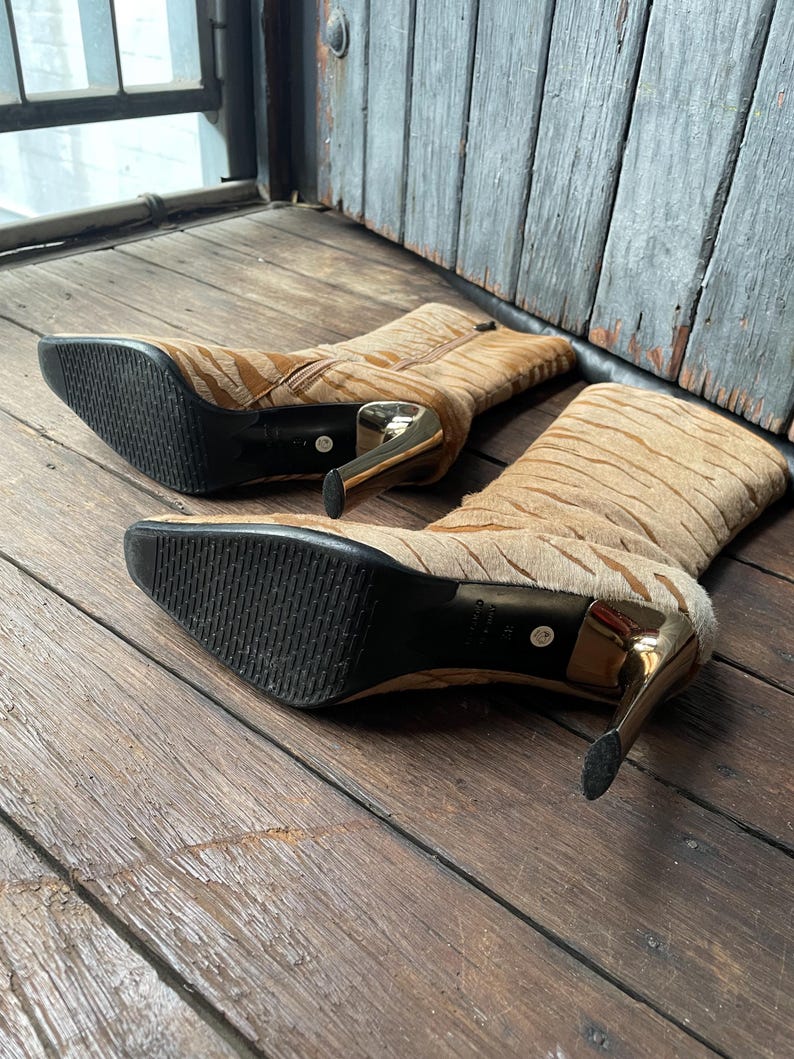 May include: A pair of tan and brown patterned high-heeled boots with gold-colored heels. The boots have a pointed toe and a zipper closure. The soles are black. The boots are made of a textured material.