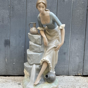 Puede incluir: Una figura de porcelana de una persona con cabello castaño claro, vistiendo una blusa azul y una falda larga blanca, apoyada sobre una pila de piedras grises. Una jarra pequeña descansa sobre las piedras y una vasija gris está en la base.