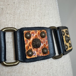 May include: A black leather belt featuring rectangular segments with textured orange and tan snakeskin-like inlays. Each segment is adorned with four smooth brass studs and a central ornate circular brass medallion, connected by brass rings.
