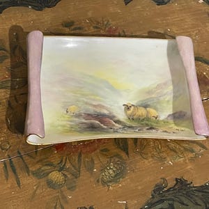 May include: A rectangular porcelain dish with a landscape painting of sheep grazing in a field. The dish has a pale pink rolled edge and a gold trim. The painting features a soft colour palette of greens, yellows, and browns.
