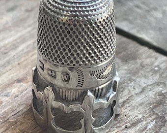 May include: A silver thimble with a textured top and a decorative, openwork design around the base. The thimble has a series of small indentations covering the main body. The thimble is made of metal and is likely used for sewing.