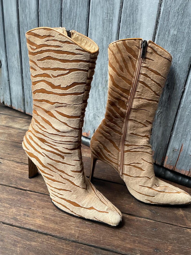 May include: A pair of tan and brown tiger-striped boots with a pointed toe and high heel. One boot has a zipper on the side. The boots are made of a textured material and are set against a wooden background.