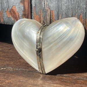 May include: A heart-shaped, iridescent white shell box with a silver-coloured metal clasp. The shell has a pearlescent sheen. The box is closed and appears to be empty, set against a weathered wooden surface.
