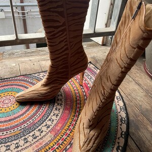 May include: A pair of brown and tan high-heeled boots with a tiger stripe pattern. The boots have pointed toes and zippers on the sides. The heels are a light brown color. The boots are on a colorful patterned rug.