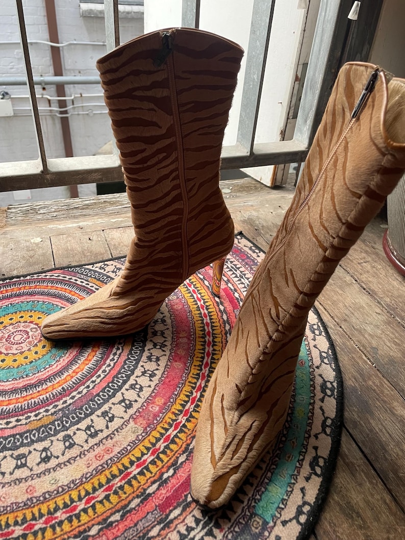 May include: A pair of brown and tan high-heeled boots with a tiger stripe pattern. The boots have pointed toes and zippers on the sides. The heels are a light brown color. The boots are on a colorful patterned rug.