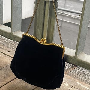 May include: A black velvet handbag with a gold-colored frame and chain strap. The purse has a curved, scalloped top and a clasp closure. The bag is hanging from a metal railing against a blurred background.