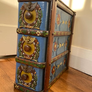 Pode incluir: Uma cômoda de madeira azul com três gavetas. Cada gaveta tem um centro amarelo com um desenho de querubim e coroa de louros. As laterais e a parte superior são decoradas com padrões florais e acabamentos.