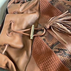 Puede incluir: Un bolso tipo saco de cuero marrón con cierre de cordón, un pasador de metal dorado, una borla larga de cuero y paneles de cuero con textura de serpiente y ante. El bolso incluye una correa para el hombro y secciones de cuero liso.