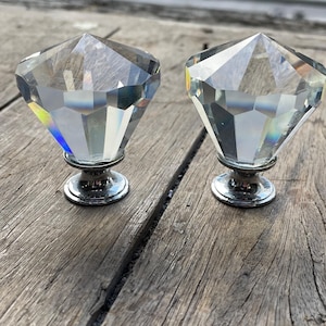 May include: Two clear, faceted crystal cabinet knobs in a diamond shape. Each knob sits on a silver-coloured base. The knobs are displayed on a weathered, grey wooden surface.