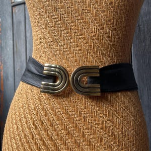 May include: A black leather belt with a wide band and a gold-colored, double-arched buckle. The belt is displayed on a mannequin with a woven, tan-colored surface. The buckle has a geometric design.