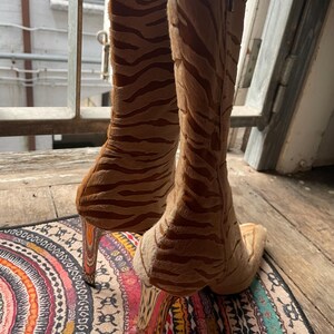 May include: A pair of high-heeled boots with a tiger stripe pattern in shades of brown and tan. The boots have a pointed toe and a tall, slender heel. The boots are made of a soft, textured material.