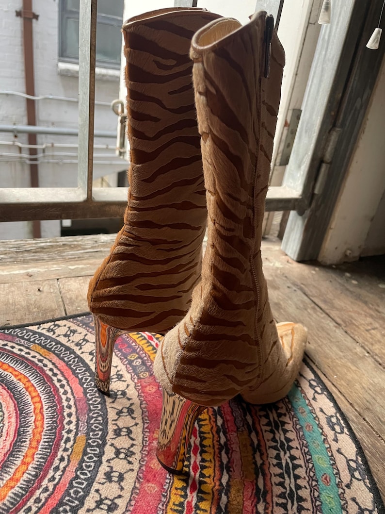 May include: A pair of high-heeled boots with a tiger stripe pattern in shades of brown and tan. The boots have a pointed toe and a tall, slender heel. The boots are made of a soft, textured material.