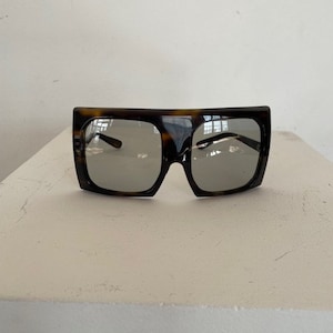May include: Square-framed sunglasses with a tortoiseshell pattern. The lenses are a reflective silver color. The sunglasses have a thick frame and a bold, modern design. The glasses are sitting on a white surface.