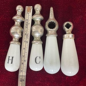 May include: Four antique faucet handles with white porcelain teardrop-shaped grips and silver-toned metal tops. Two handles are marked with "H" and "C". A ruler shows measurements in inches.
