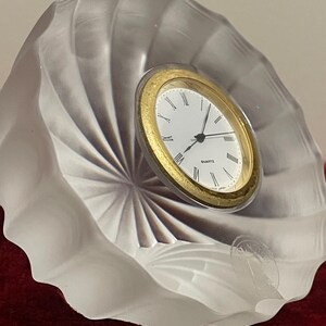 Puede incluir: Un reloj de cuarzo con bisel dorado y esfera blanca, con números romanos y manecillas negras. El reloj está dentro de una escultura de vidrio esmerilado y arremolinado. La base es de color rojo oscuro.