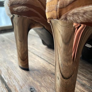 May include: Close-up of two high-heeled shoes. The heels are a metallic gold color with a wood grain pattern. The uppers are a tan and brown zebra print. The shoes are on a wooden surface.