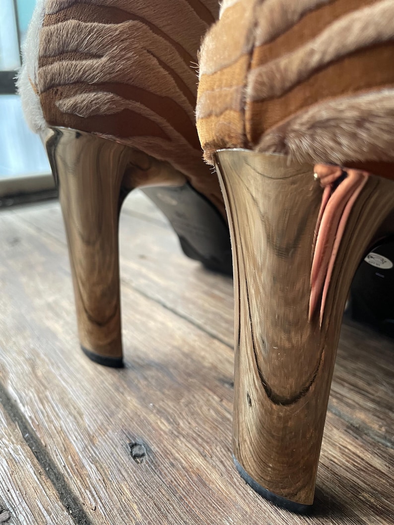 May include: Close-up of two high-heeled shoes. The heels are a metallic gold color with a wood grain pattern. The uppers are a tan and brown zebra print. The shoes are on a wooden surface.