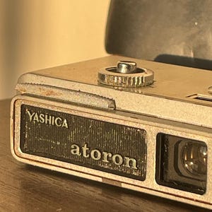 以下が含まれることがあります： シルバーボディのヴィンテージYashica Atoronカメラ。カメラは、顕著なレンズと上部のテクスチャードダイヤルを備えています。「Yashica」と「atoron」という名前がカメラの前面に印刷されています。