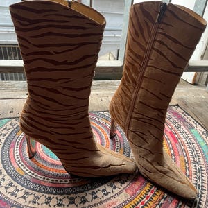 May include: A pair of brown and tan high-heeled boots with a tiger stripe pattern. The boots have a pointed toe and a zipper on the side. The heels are clear. The boots are on a colorful patterned rug.
