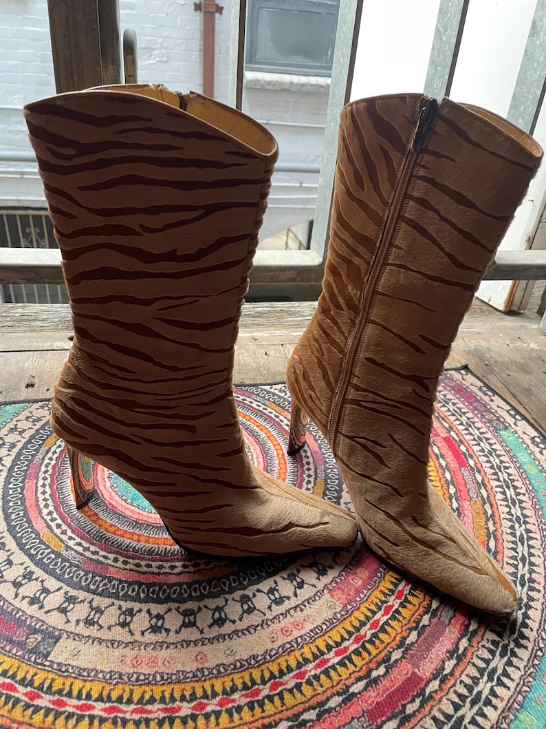 May include: A pair of brown and tan high-heeled boots with a tiger stripe pattern. The boots have a pointed toe and a zipper on the side. The heels are clear. The boots are on a colorful patterned rug.