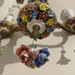 May include: Close-up of a vintage chandelier with floral accents. The chandelier features a white base with colorful ceramic flowers in shades of pink, blue, yellow, and purple. The metal arms and details have a gold finish. The light fixtures are visible.