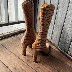 May include: A pair of knee-high boots with a tiger stripe pattern in shades of tan and brown. The boots have a high heel and a zipper closure. The boots are made of a soft, textured material.