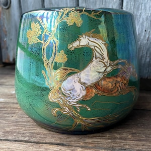 May include: A round, iridescent green vase with a detailed illustration of a white horse rearing up near a gold-coloured tree. The vase has a glossy finish and is set against a wooden surface.
