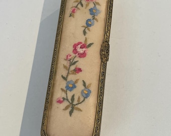 May include: An antique rectangular compact with a floral embroidered design. The compact features a cream-colored fabric with pink and blue flowers, set within a gold-toned metal frame. The frame has an ornate, decorative edge and a clasp closure.