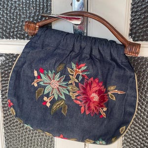 May include: A dark blue handbag with a wooden handle and floral embroidery. The bag features a curved wooden handle and is adorned with embroidered flowers in shades of red, green, and teal. The bag has a beige trim.