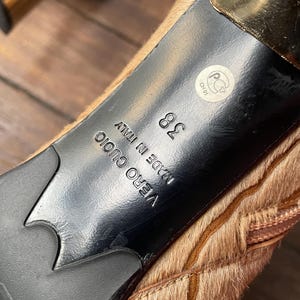 May include: Close-up of the sole of a high-heeled shoe. The sole is black with a gold heel and a light brown, textured upper. The shoe size is 38, and the words "VERO CUOIO MADE IN ITALY" are visible.