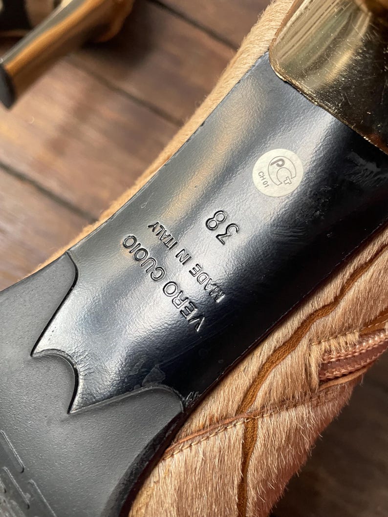 May include: Close-up of the sole of a high-heeled shoe. The sole is black with a gold heel and a light brown, textured upper. The shoe size is 38, and the words "VERO CUOIO MADE IN ITALY" are visible.