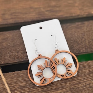 May include: A pair of wooden earrings with a sun design. The earrings are round and have a silver hook.