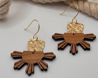 May include: A pair of wooden earrings with a gold accent. The earrings are shaped like a sun with rays coming out from the center. The gold accent is a small, textured piece of metal.