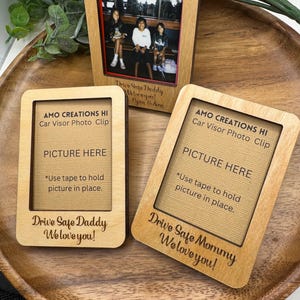 May include: Two wooden photo frames with a brown background. The frames are designed for car visors and have the text "AMO CREATIONS HI Car Visor Photo Clip" and "Drive Safe Daddy We love you!" and "Drive Safe Mommy We love you!" The frames have a space for a picture and instructions to use tape to hold the picture in place.