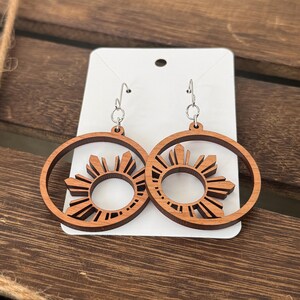 May include: A pair of wooden earrings with a sunburst design. The earrings are brown and have a silver hook.