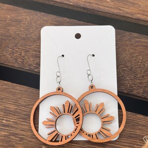 May include: A pair of wooden earrings with a sun design. The earrings are brown and have a silver hook.